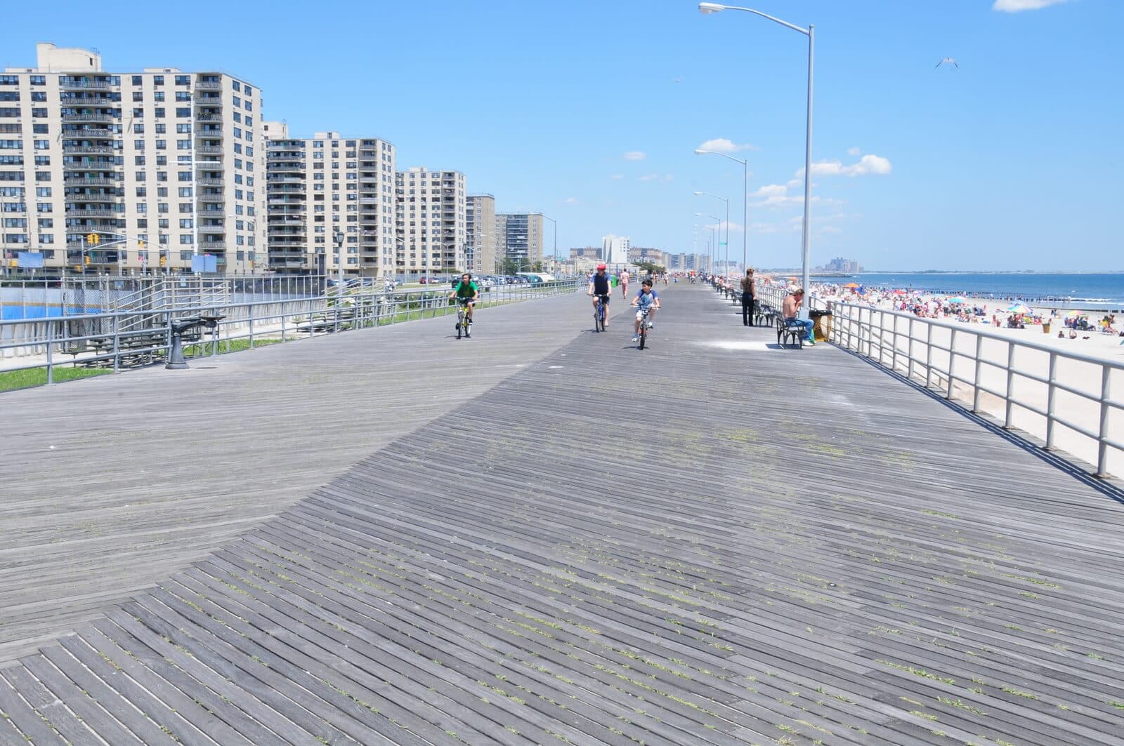 Wide wooden boardwalk by the beach with people walking and biking, high-rise buildings on the left, and a busy sandy shoreline on the right under a clear blue sky