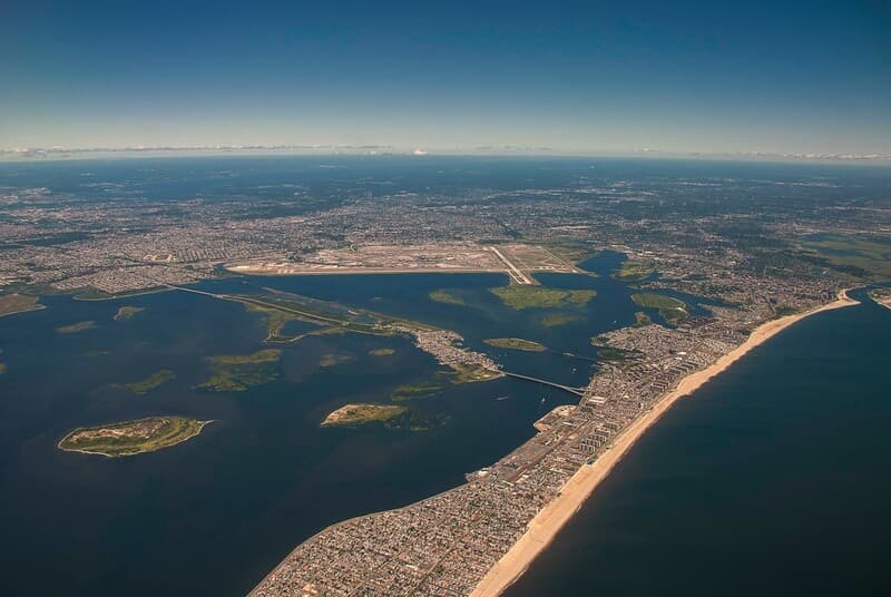 Aerial view of Rockaway Beach, NY, with bridges, waterways, and an airport visible in the background under a clear sky.